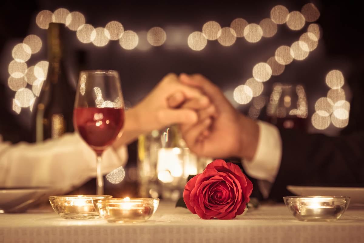 coppia che si dà la mano a tavola con una rosa rossa e un calice di vino