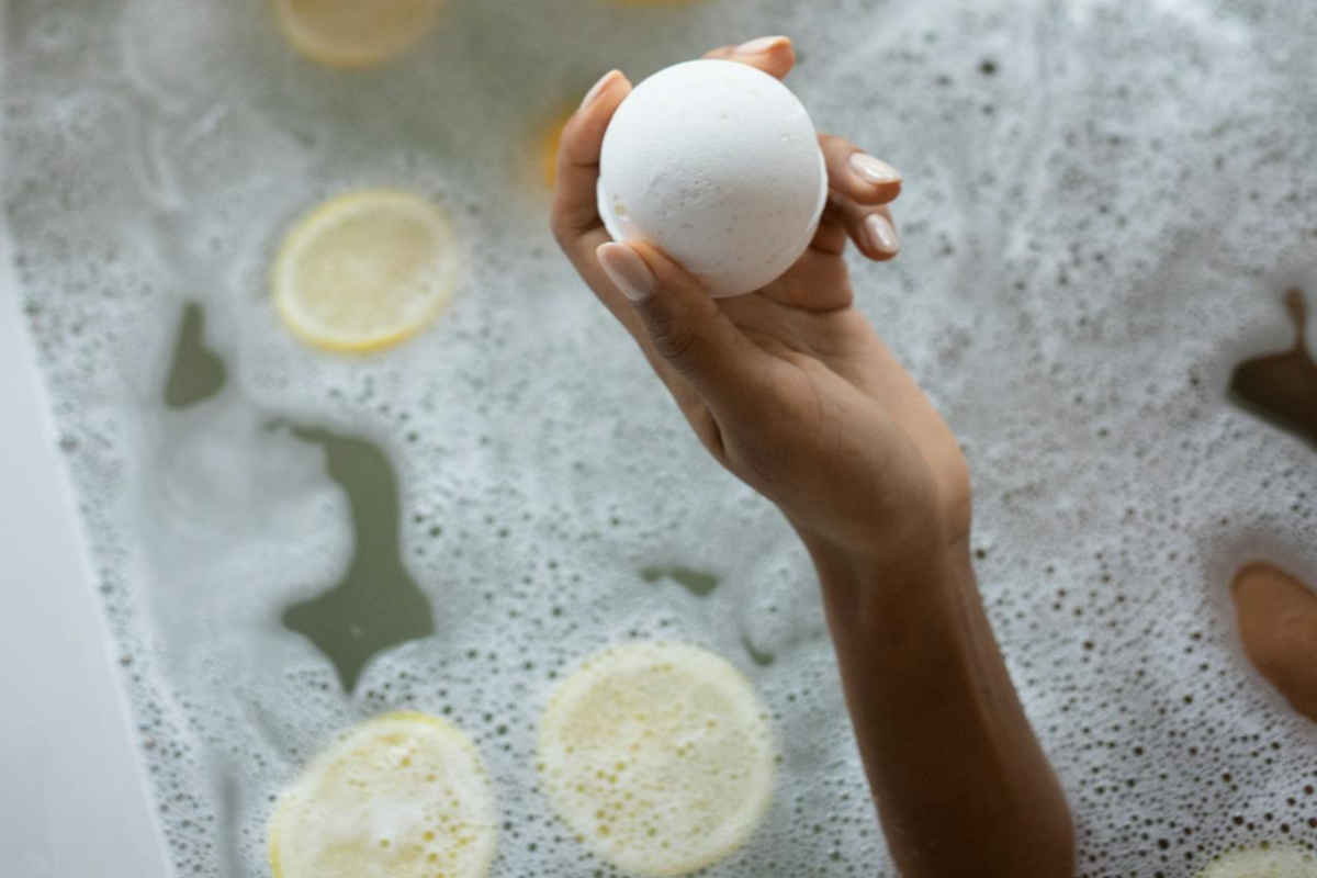 Vasca da bagno con acqua, limoni e una mano che tiene una bomba da bagno